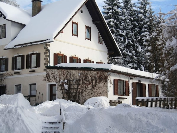 A two-story building with a sloped roof covered in snow, surrounded by snow-covered trees and a wooden fence.