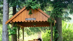 Um gazebo de madeira com uma placa, rodeado por árvores, apresentando uma churrasqueira e mesas de piquenique.