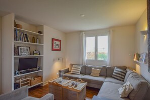 A living room with a sofa, a television on a stand, a bookshelf, and a window with curtains. - Holiday Home in Dordogne Estate (Saint-Medard-d'Excideuil)