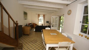 A dining area with a wooden table and white chairs, a staircase, a patterned floor, and a view of the outdoors through a window. - Holiday Home in Ardeche Near Chateau Ventadour (Lalevade-d'Ardeche)