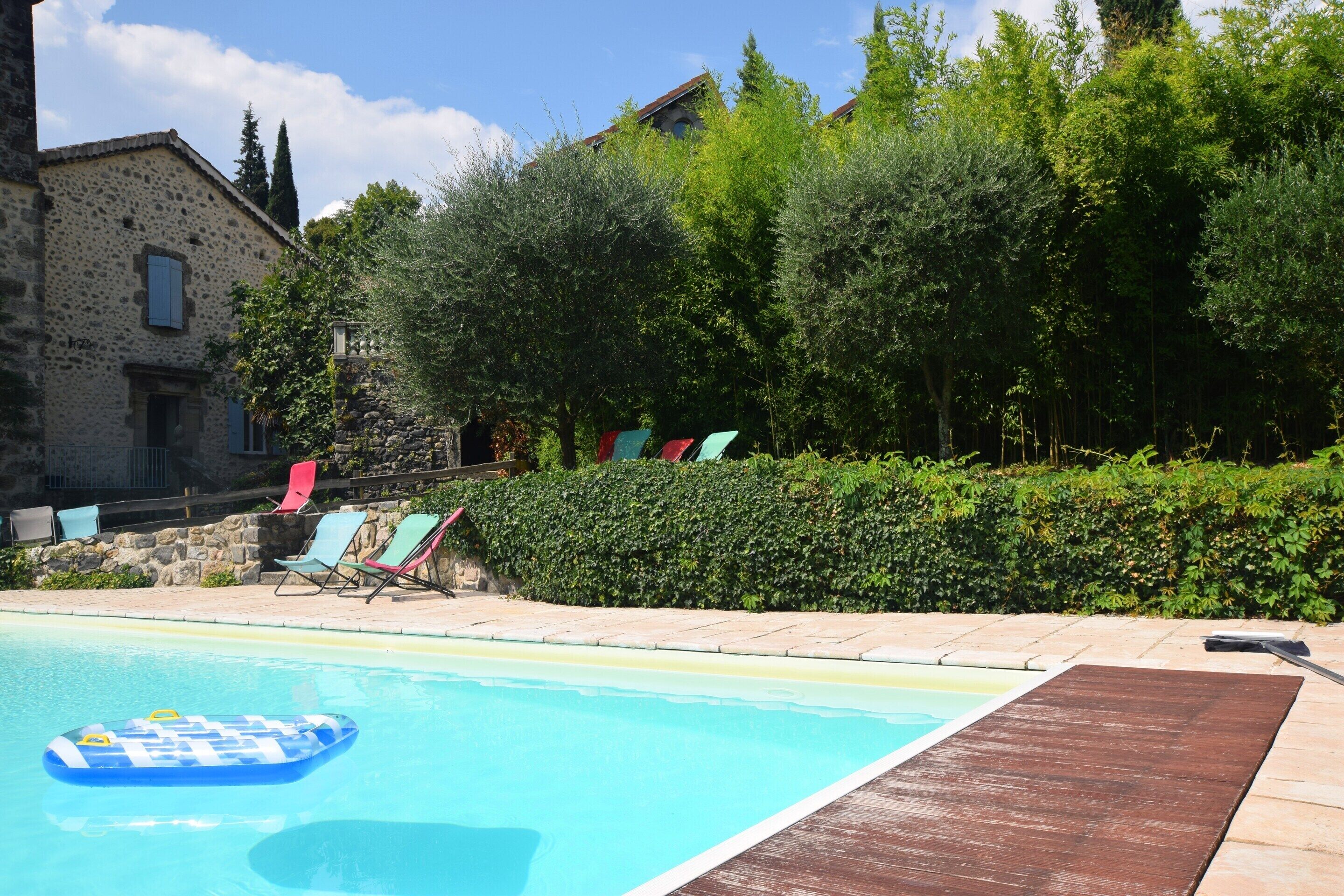 A swimming pool with a wooden deck, surrounded by greenery and a stone wall.