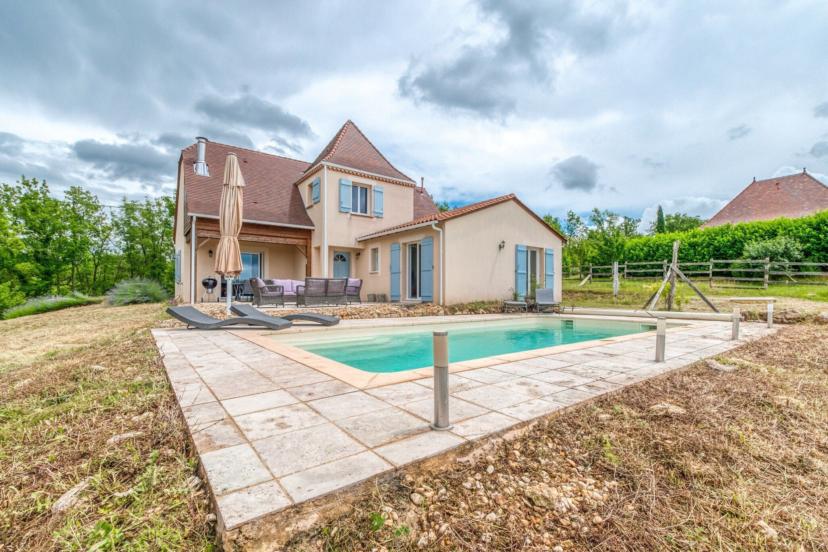Una casa de dos pisos con piscina, mobiliario exterior y jardín cercado.