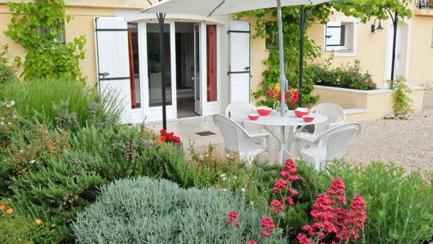Un espace extérieur aménagé avec des chaises blanches et une table, entouré de différentes plantes et fleurs.