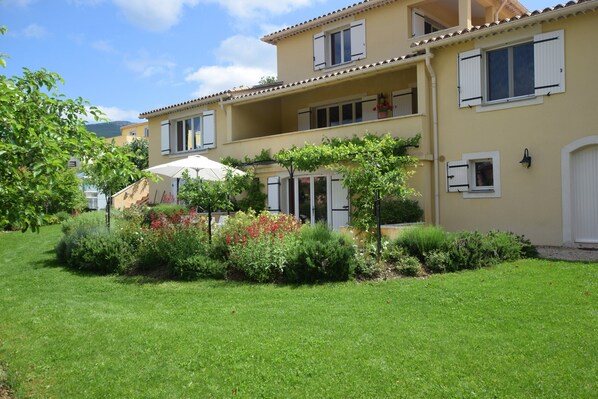A two-story building with a balcony, a garden with various plants, and a white door. - Modern Terrace Stay Montbrun (Montbrun-les-Bains)