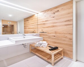 A modern bathroom with a wooden panel wall, a large mirror, a white sink, and a wooden shelf with towels and a hairdryer.