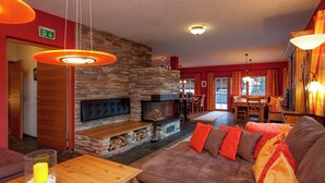 A cozy living room with a stone fireplace, a wooden coffee table, and a brown sofa with orange cushions. - Posh Mansion in Sankt with 2 Saunas, Whirlpool, Hot Tub (Sankt Anton am Arlberg)