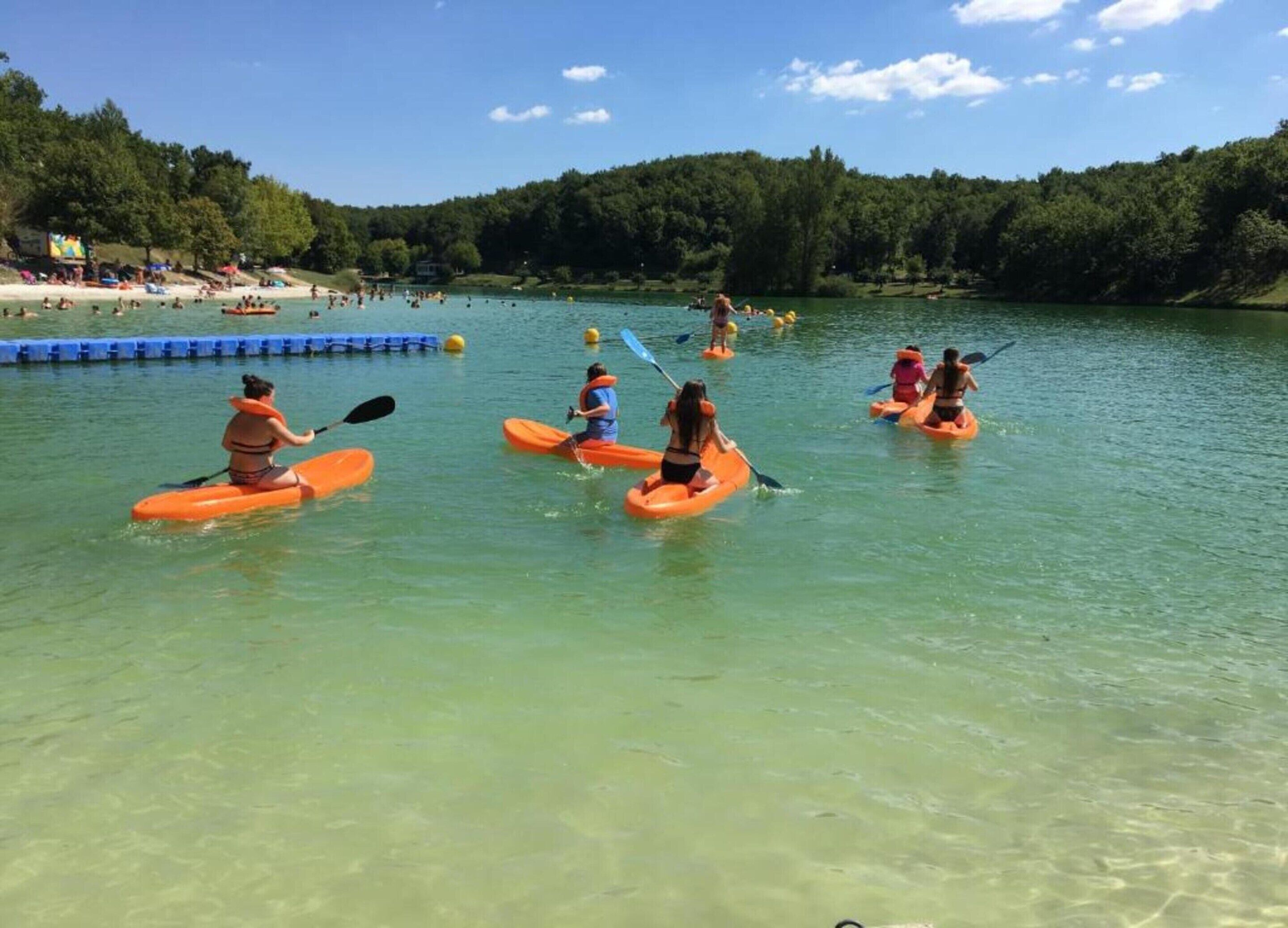 People kayaking on a calm lake with clear water and a forested hillside in the background.