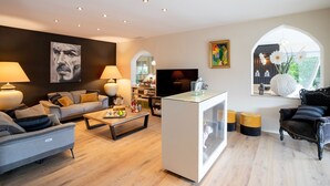 Ein modernes Wohnzimmer mit einer Couch, einem Couchtisch und einem Fernseher. Es gibt Lampen, ein Gemälde und ein Fenster mit Blick.