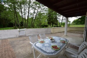 Une terrasse extérieure avec une table dressée pour deux personnes, entourée de chaises blanches, et une vue sur une zone boisée.