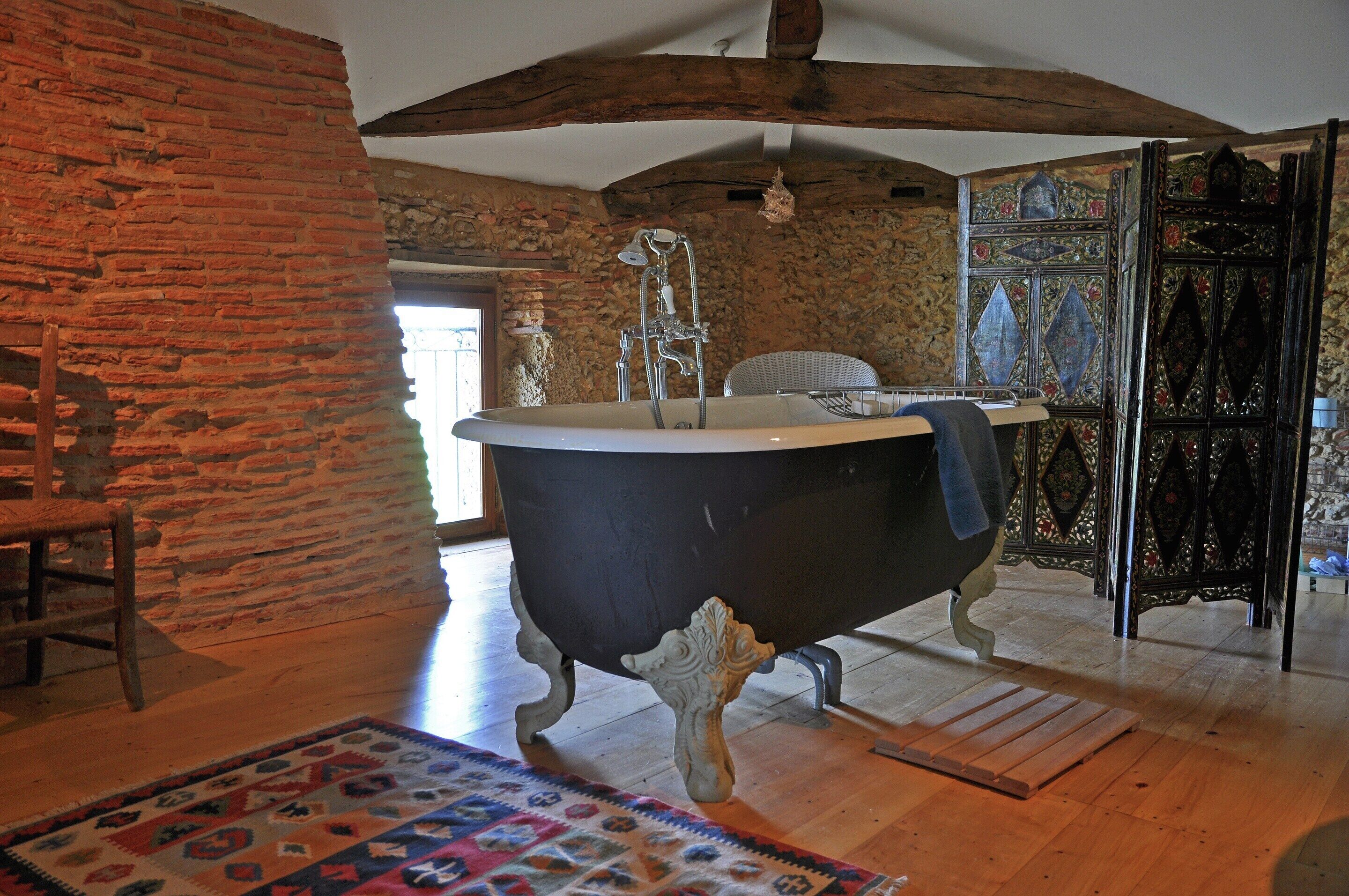 Una habitación con una bañera de patas de león, una silla de madera, un tapete estampado y una pared de piedra.