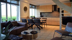 A modern living room with a sofa, chairs, and a coffee table. There is a kitchen area with wooden cabinets and a dining table. - Dike House in Schoondijke Near Beaches (Schoondijke)