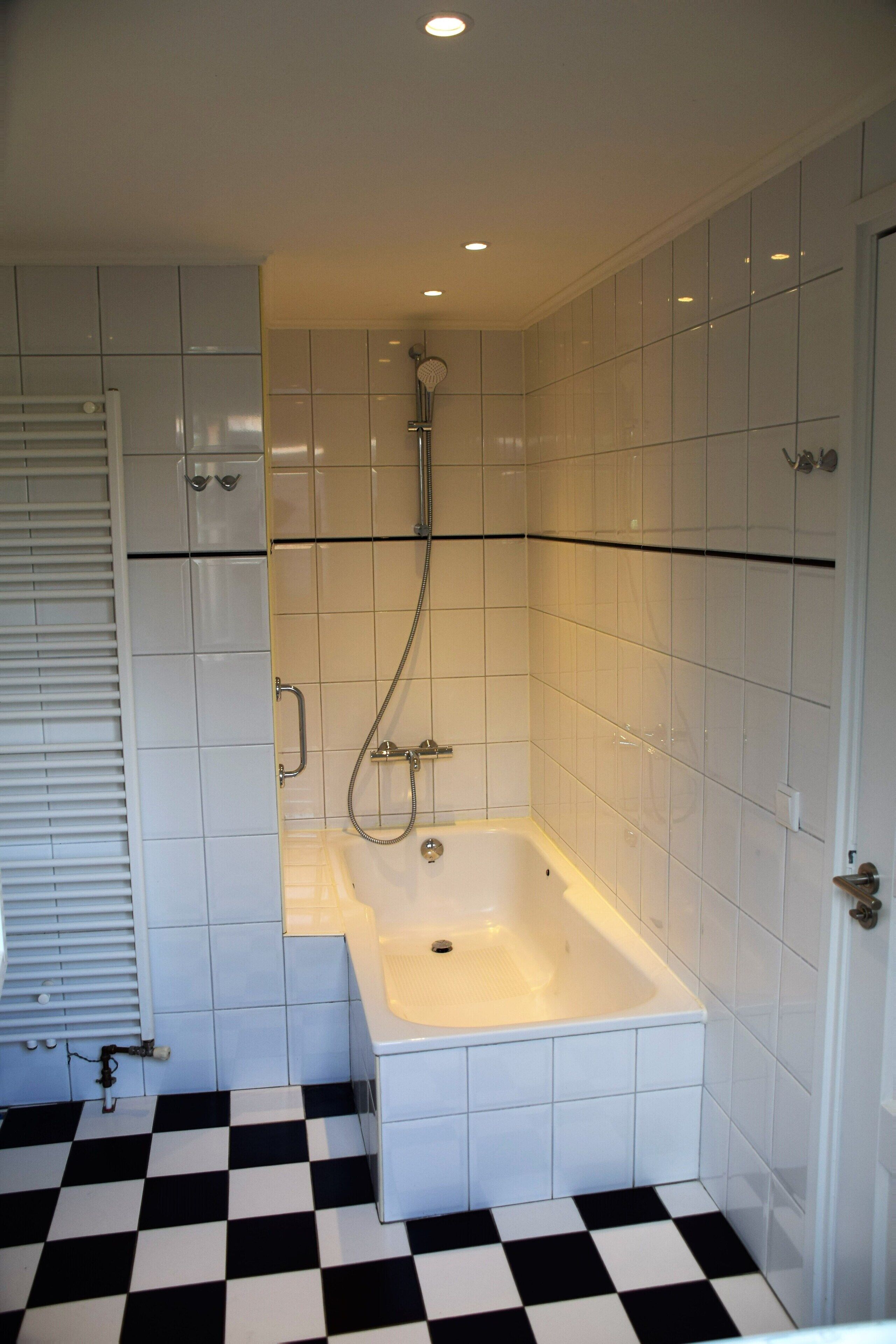 Un baño con azulejos blancos, suelo a cuadros blanco y negro, una bañera y una ducha.