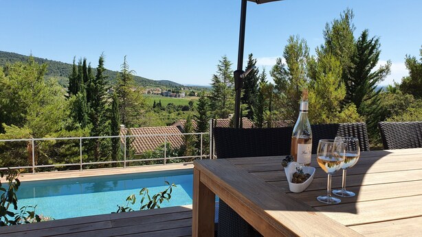 Una terraza de madera con piscina, una botella de vino y vasos sobre una mesa, con vistas a la piscina y un paisaje pintoresco.