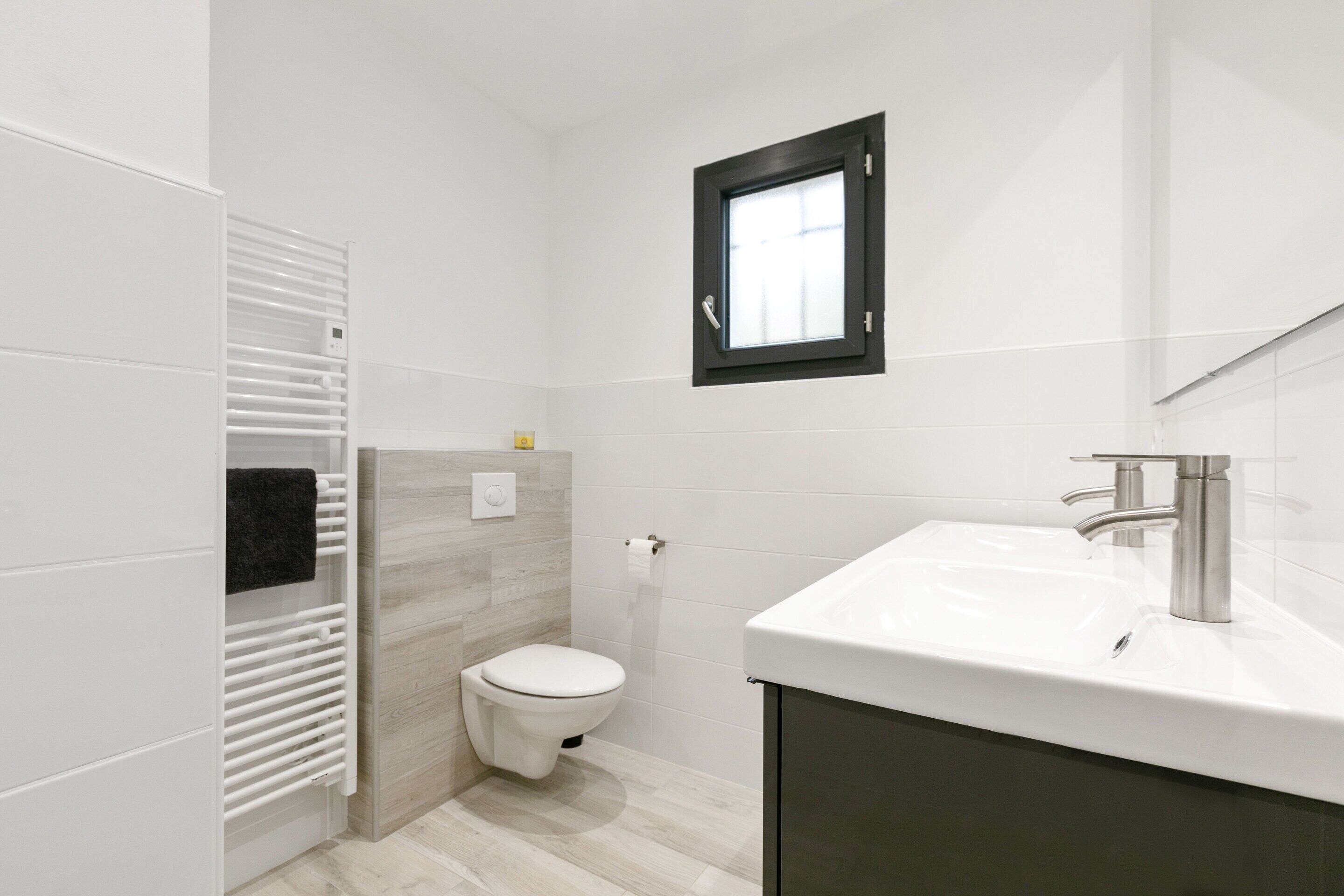 Une salle de bain moderne avec des toilettes blanches, un lavabo blanc doté d’un robinet argenté et une petite fenêtre située au-dessus des toilettes.