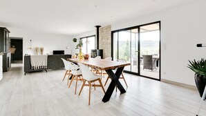 Un comedor amplio con una gran mesa de madera y sillas blancas, una zona de cocina con un muro de acento de piedra, y un toldo de vidrio corredizo que da a un patio exterior.