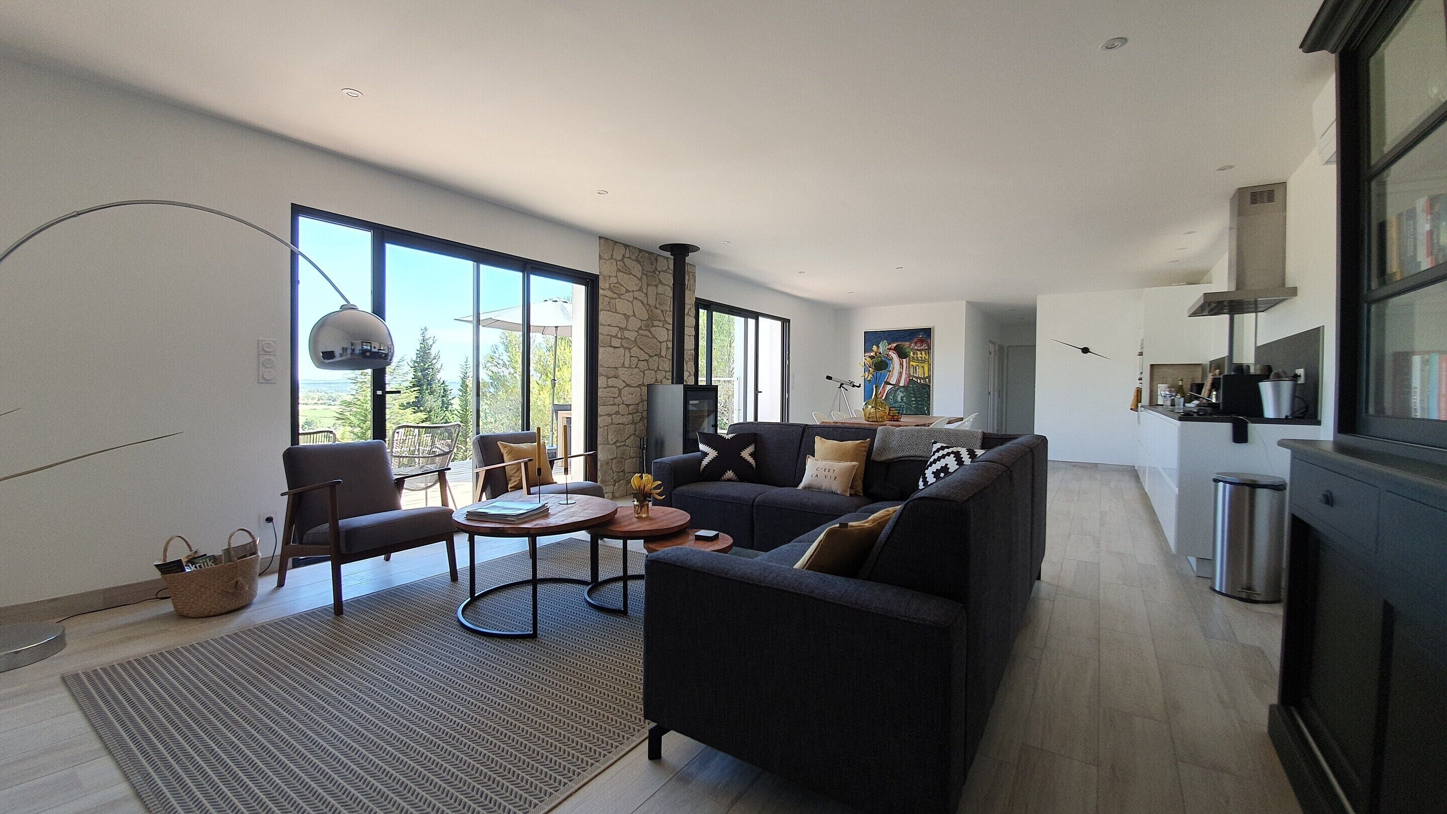 A modern living room with a sofa, coffee table, and chairs, a kitchen area with a stove and cabinets, and large windows offering a view of the outdoors.