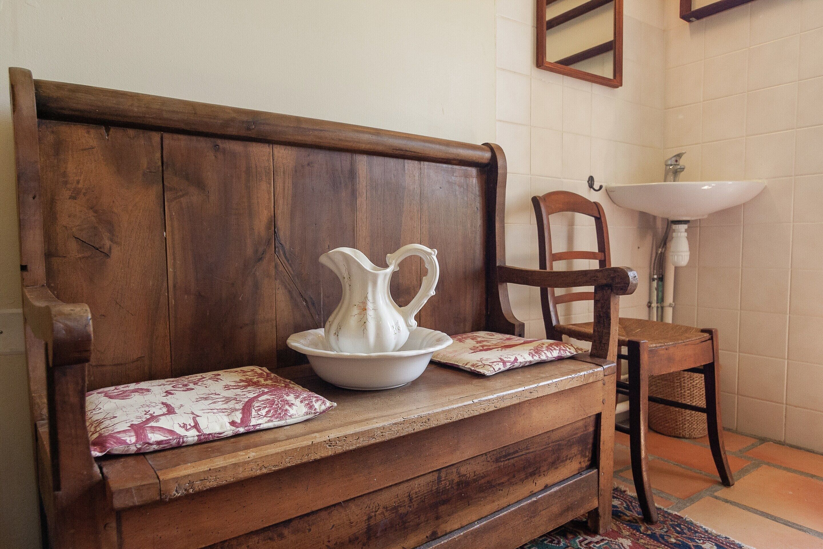 A wooden bench with a white pitcher and a cushion, a wooden chair, and a white sink in a room with tiled walls.