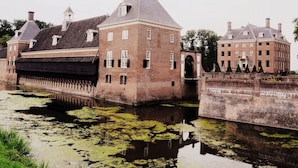 A historic building with a large brick structure, a water canal in front, and a bridge connecting to another building. - Peaceful Forest Chalet (Elst)