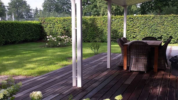 A covered outdoor seating area with a wooden deck, white pillars, and wicker furniture. - Peaceful Forest Chalet (Elst)