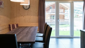 Een houten interieur met een eettafel, stoelen, een klok en uitzicht op een houten gebouw door een glazen deur.