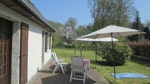 A patio with two chairs and a table, a large umbrella, and a grassy area with trees and a building in the background. - Bungalow in France Near Castle & Hiking Trails (Saint-Pierre-le-Vieux)