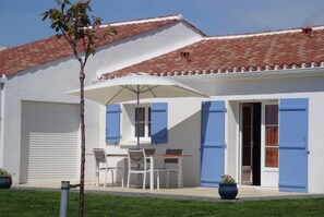 A building with a tiled roof, white walls, blue shutters, a table with chairs, and a potted plant. - Holiday Home in Avrille Near Sandy Beaches (Avrille)