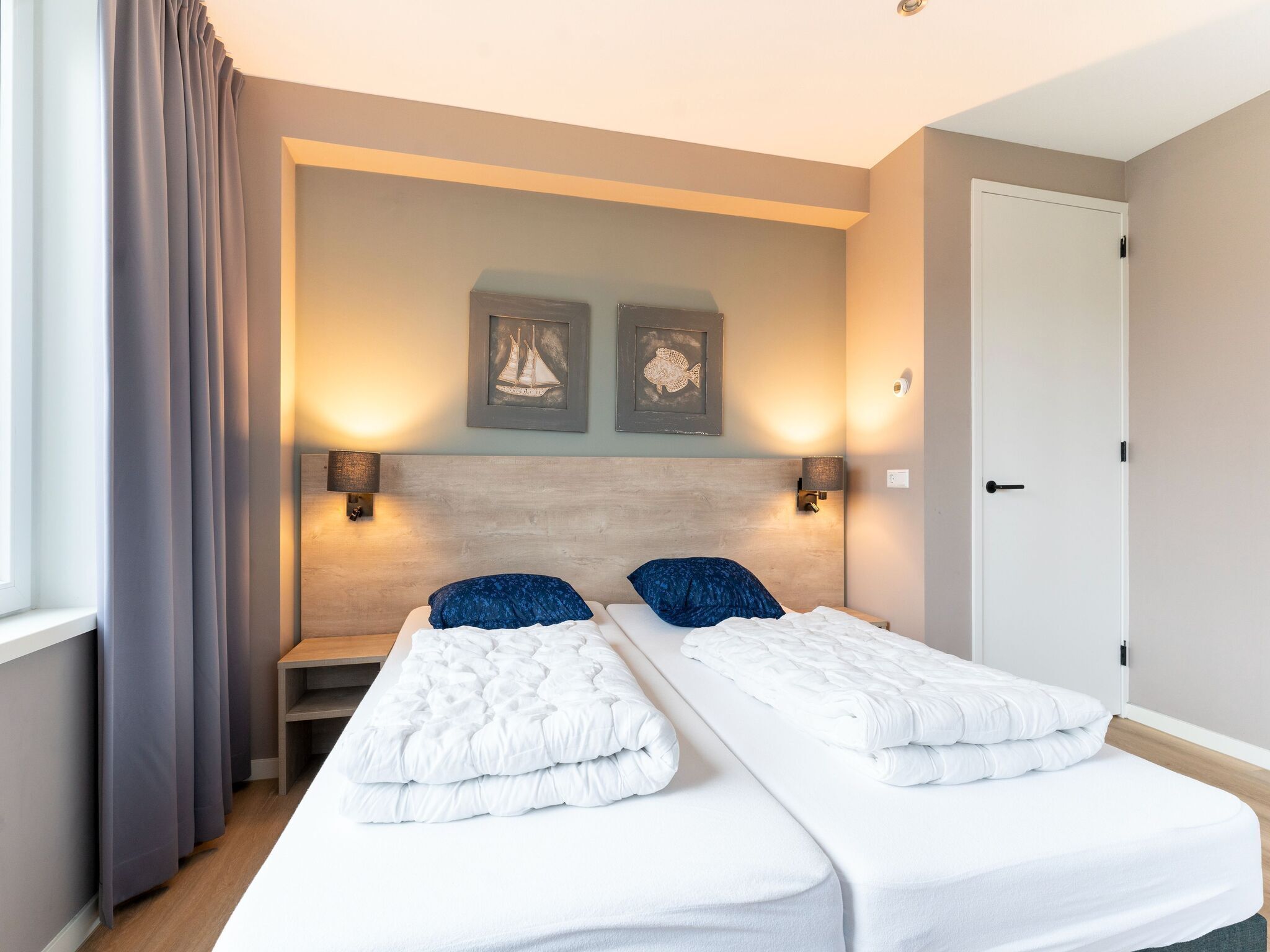 Quarto de hotel com duas camas, cabeceira de madeira, luminárias fixadas na parede e obras de arte emolduradas.