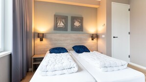 A hotel room with two beds, a wooden headboard, wall-mounted lamps, and framed artwork. - Holiday Home in Texel Near the Sea (De Koog)