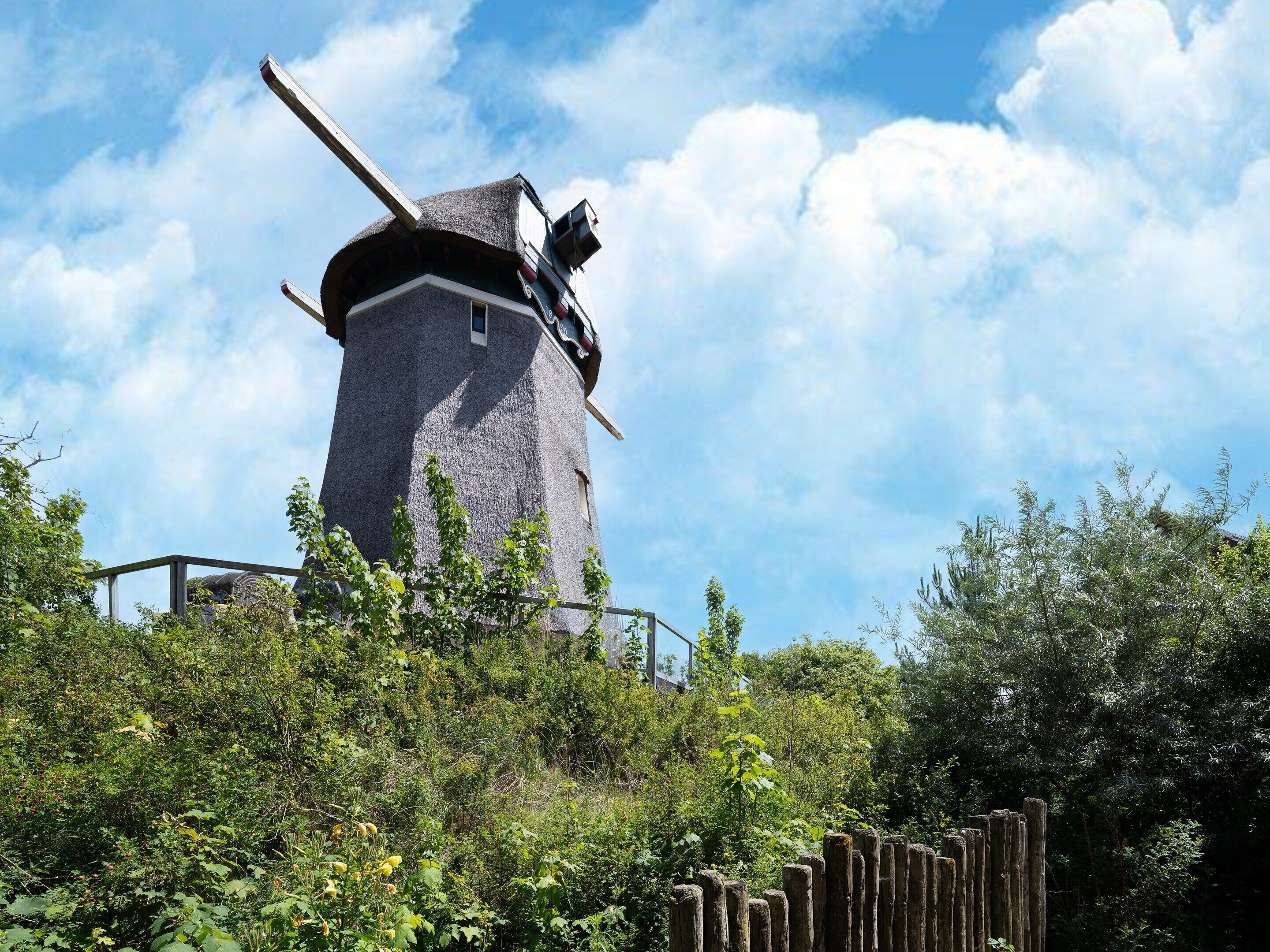 Un moulin à vent au design traditionnel, entouré d’une végétation luxuriante et sous un ciel dégagé.