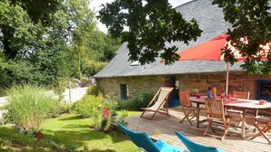 A stone building with a red roof, a wooden deck with a table and chairs, and a garden with various plants. - 17th Century Breton Cottage With Modern Interior on the Edge of a Forest (Huelgoat)