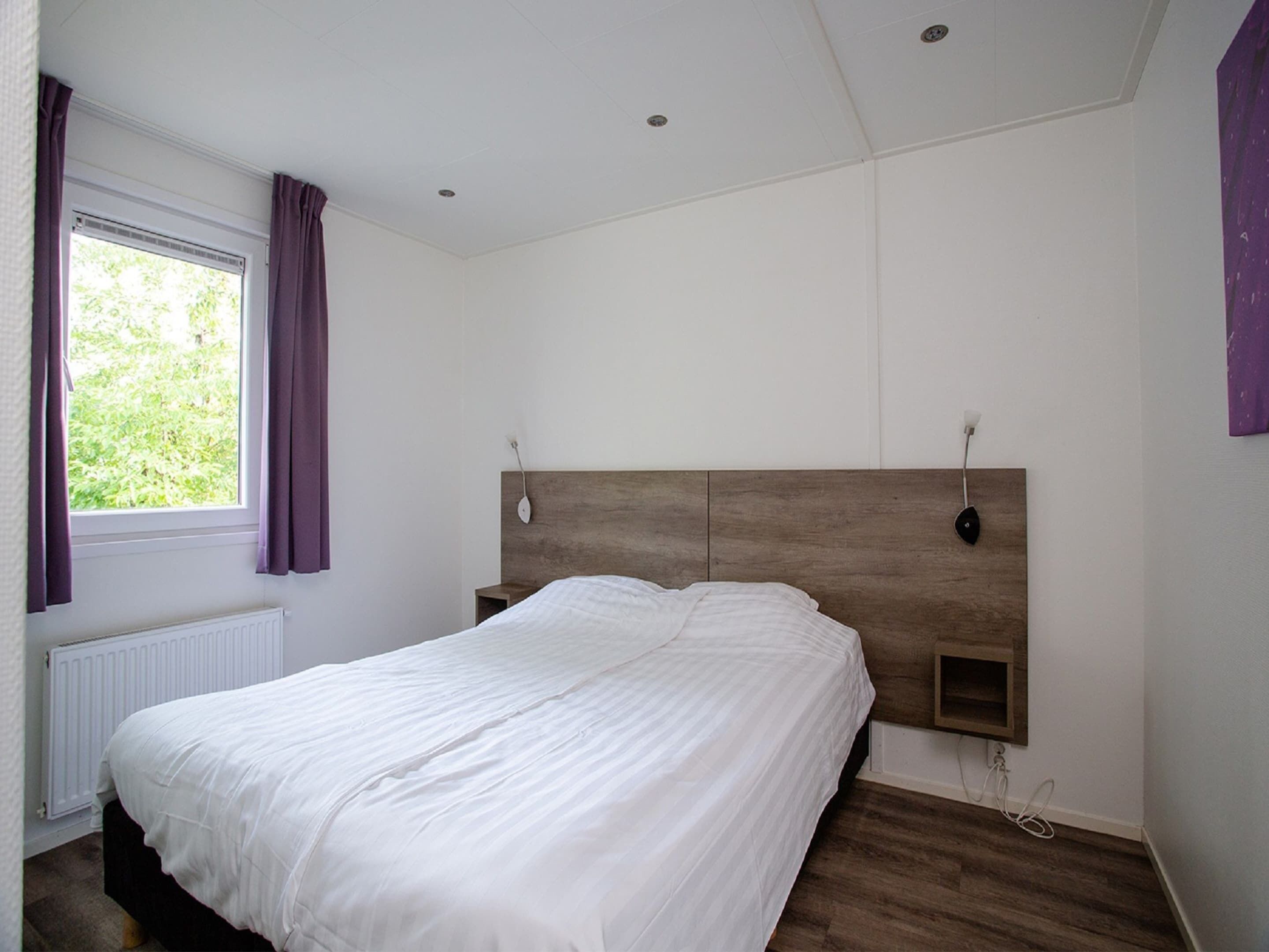 A bedroom with a wooden headboard, a white bedspread, a window with purple curtains, and a white radiator.
