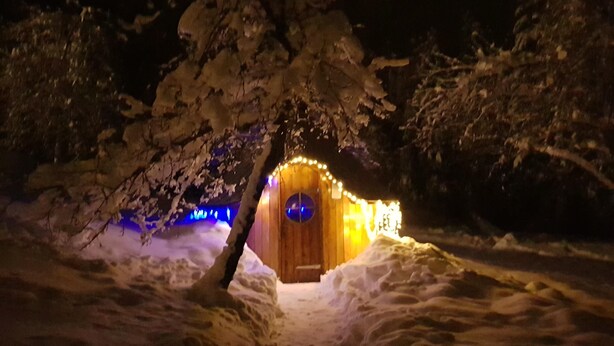 Lumipeitteinen maisema, jossa näkyy valoilla koristeltu puinen mökki.