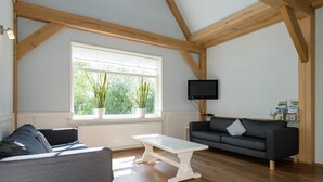 A spacious living room with a high ceiling, wooden beams, a large window with plants, a black TV, and a grey sofa. - Inviting Holiday Home in Zuidoostbeemster near Center & Forest (Zuidoostbeemster)