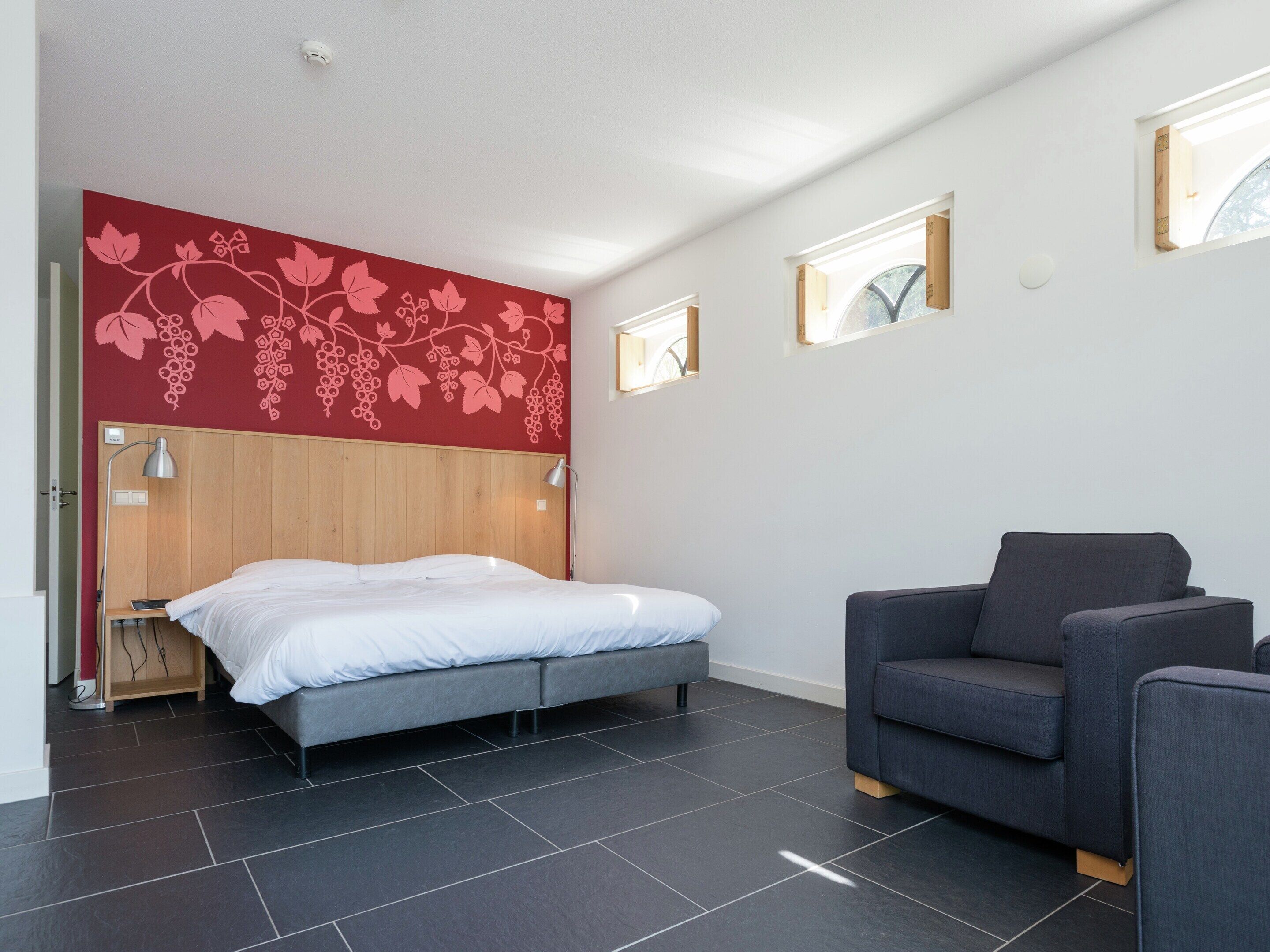 A modern bedroom with a bed, a grey sofa, and a red wall with a grapevine design.