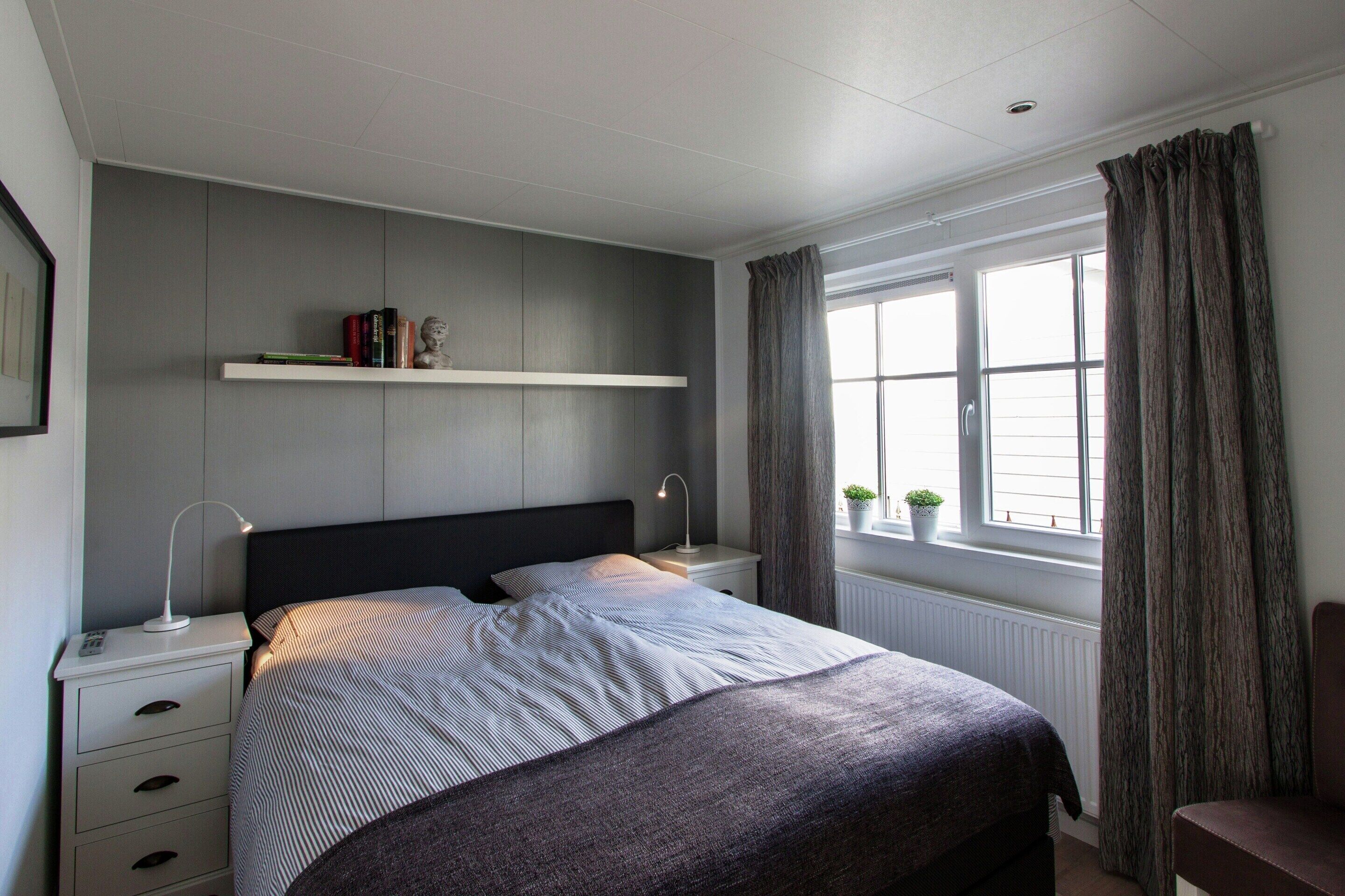 A bedroom with a bed, bedside tables, a shelf with books, a window with curtains, and a radiator.