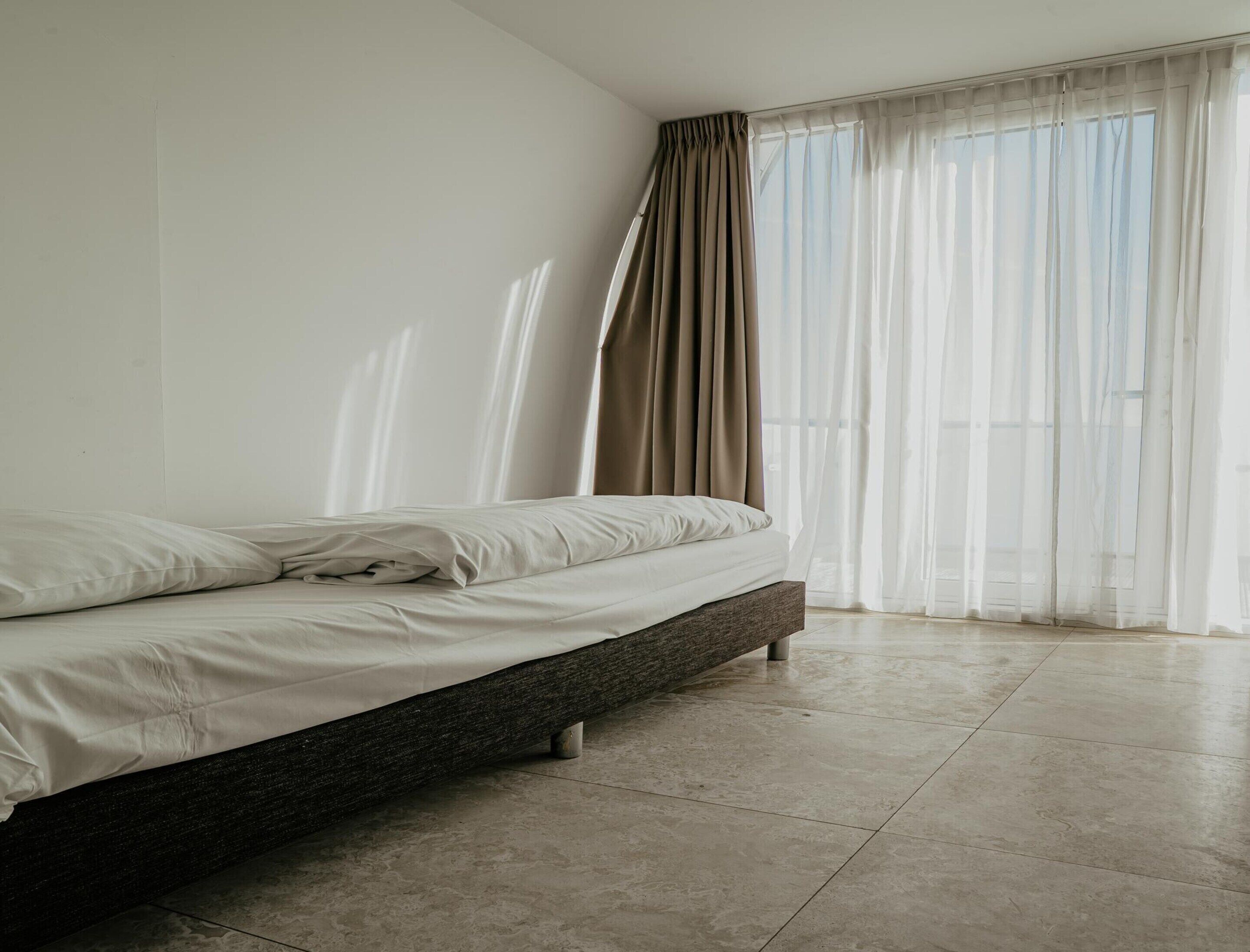 A modern bedroom with a large bed, a window with sheer curtains, and a tiled floor.