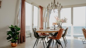 A dining area with a large table, chairs, a chandelier, and a view of the sea through large windows. - Apartment in Egmond With Sea Views (Egmond aan Zee)