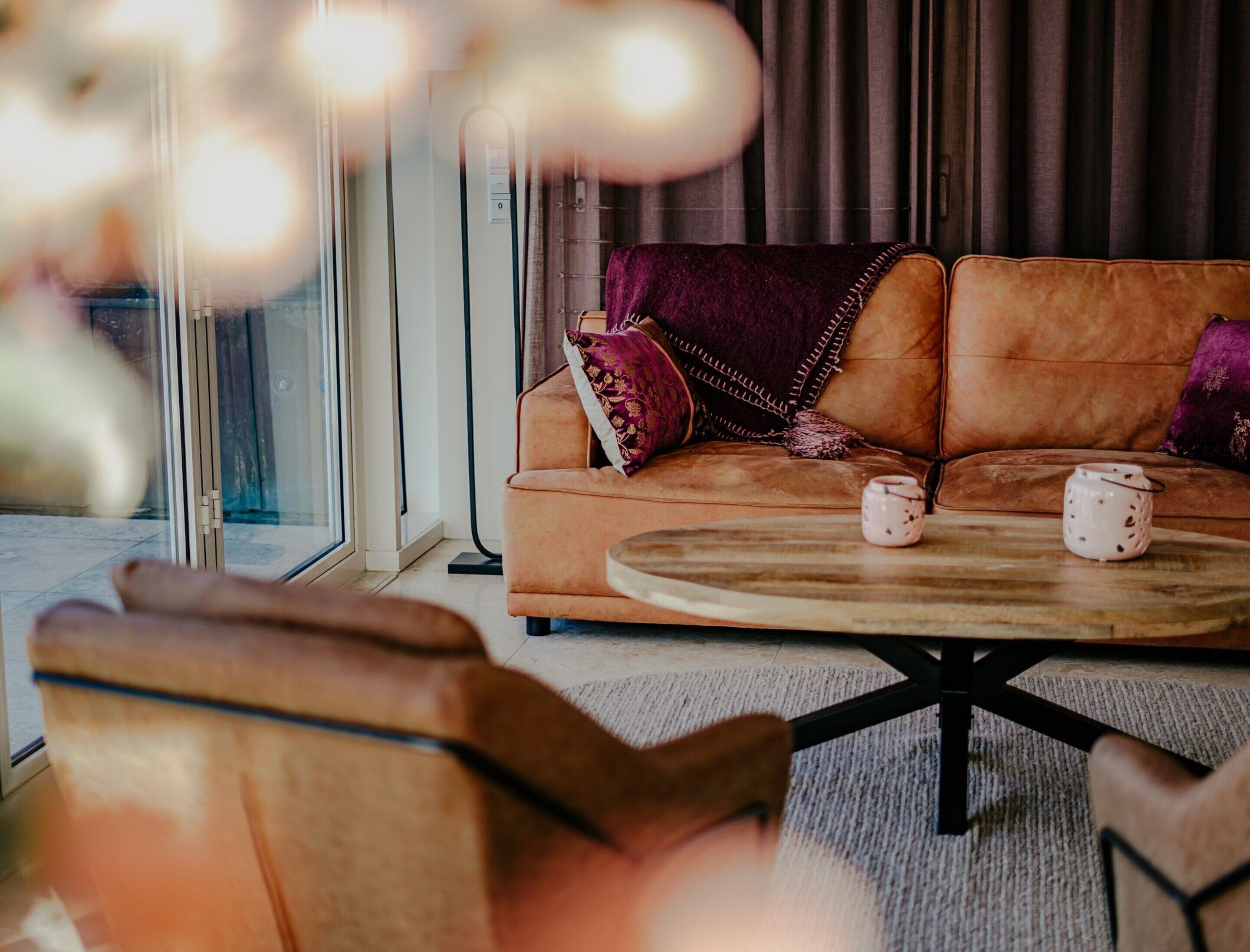 A cozy living room with a brown leather sofa, two pillows, a round wooden coffee table, and a glass door leading outside.