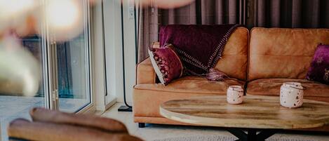 A cozy living room with a brown leather sofa, two pillows, a round wooden coffee table, and a glass door leading outside.