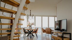 A spacious living area with a large window overlooking the sea, a dining table with chairs, a TV, and a staircase with wooden beams. - Apartment in Egmond With Sea Views (Egmond aan Zee)