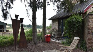 A rustic outdoor seating area with a wooden lounge chair, a red trash bin, and a brick building with a tiled roof. - Apartment Near the Belgian Border (Eijsden)