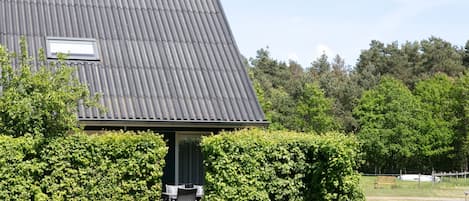 A building with a corrugated metal roof, a small window, and a door. There is a wooden fence and a hedge in front of the building.