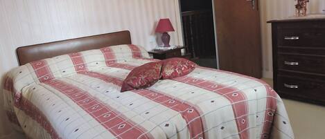 Una cama con colcha a cuadros, una mesita de noche con lámpara y un aparador con cajones.