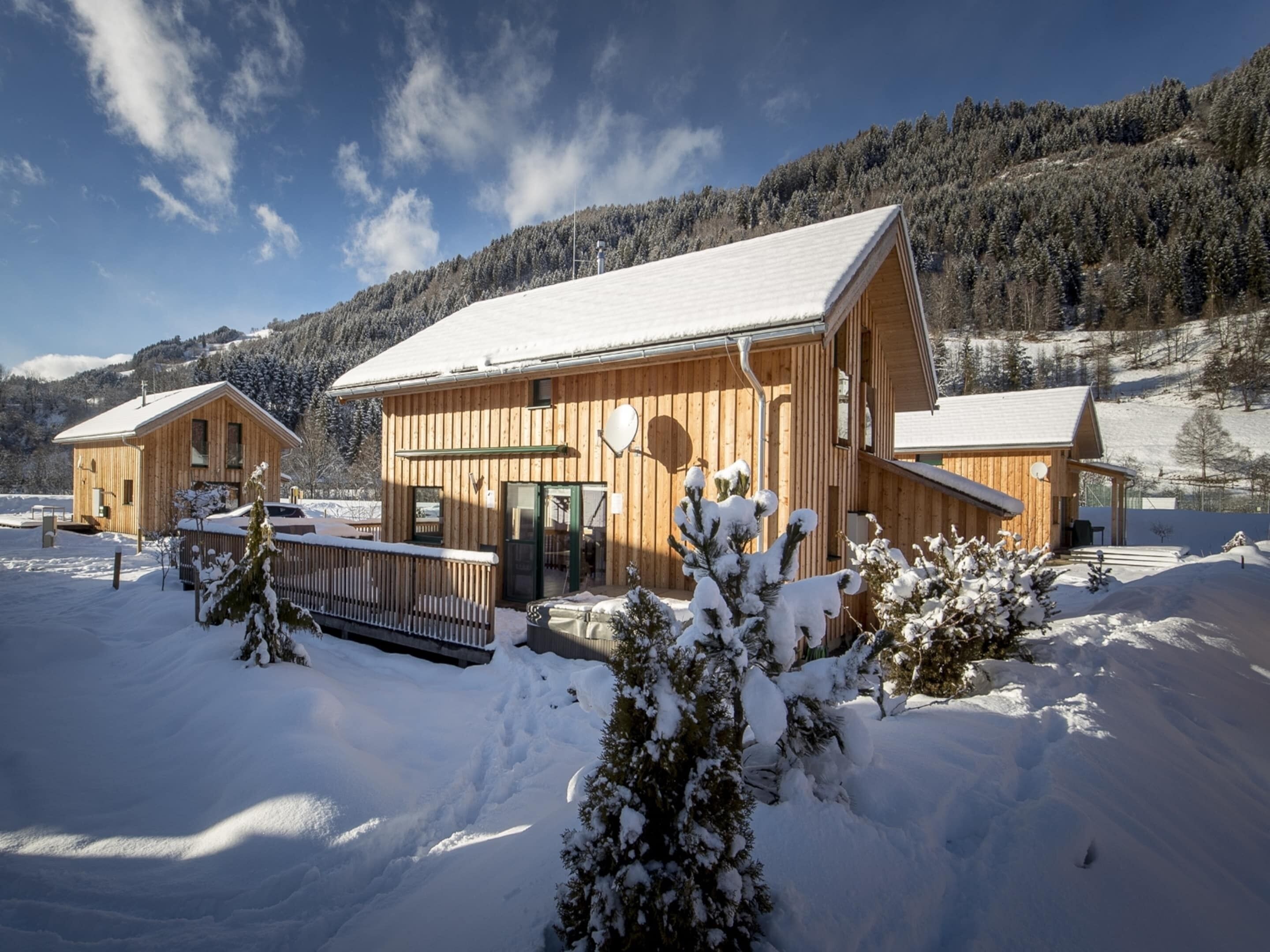 Un chalet de madera con techo nevado, rodeado de un paisaje invernal y una montaña boscosa al fondo.
