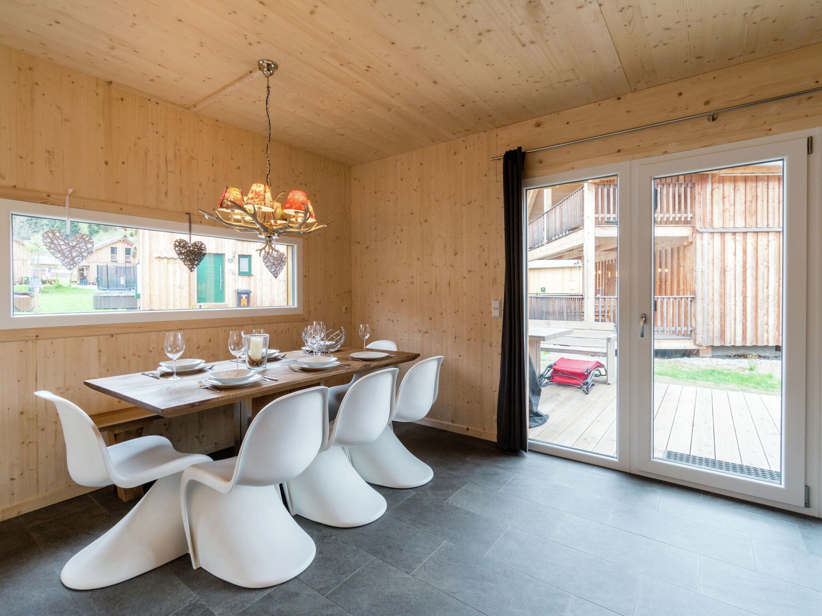 A wooden interior with a dining table set for four, white chairs, a chandelier, and a sliding glass door leading outside.