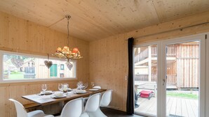 A wooden interior with a dining table set for four, white chairs, a chandelier, and a sliding glass door leading outside. - Cosy Chalet With hot tub (Sankt Georgen am Kreischberg)