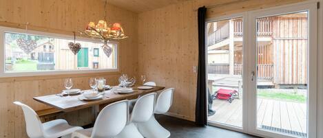 A wooden interior with a dining table set for four, white chairs, a chandelier, and a sliding glass door leading outside.