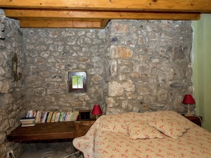 A stone-walled room with a wooden ceiling, a small window, a bookshelf, a bed with floral bedding, and two red lamps.