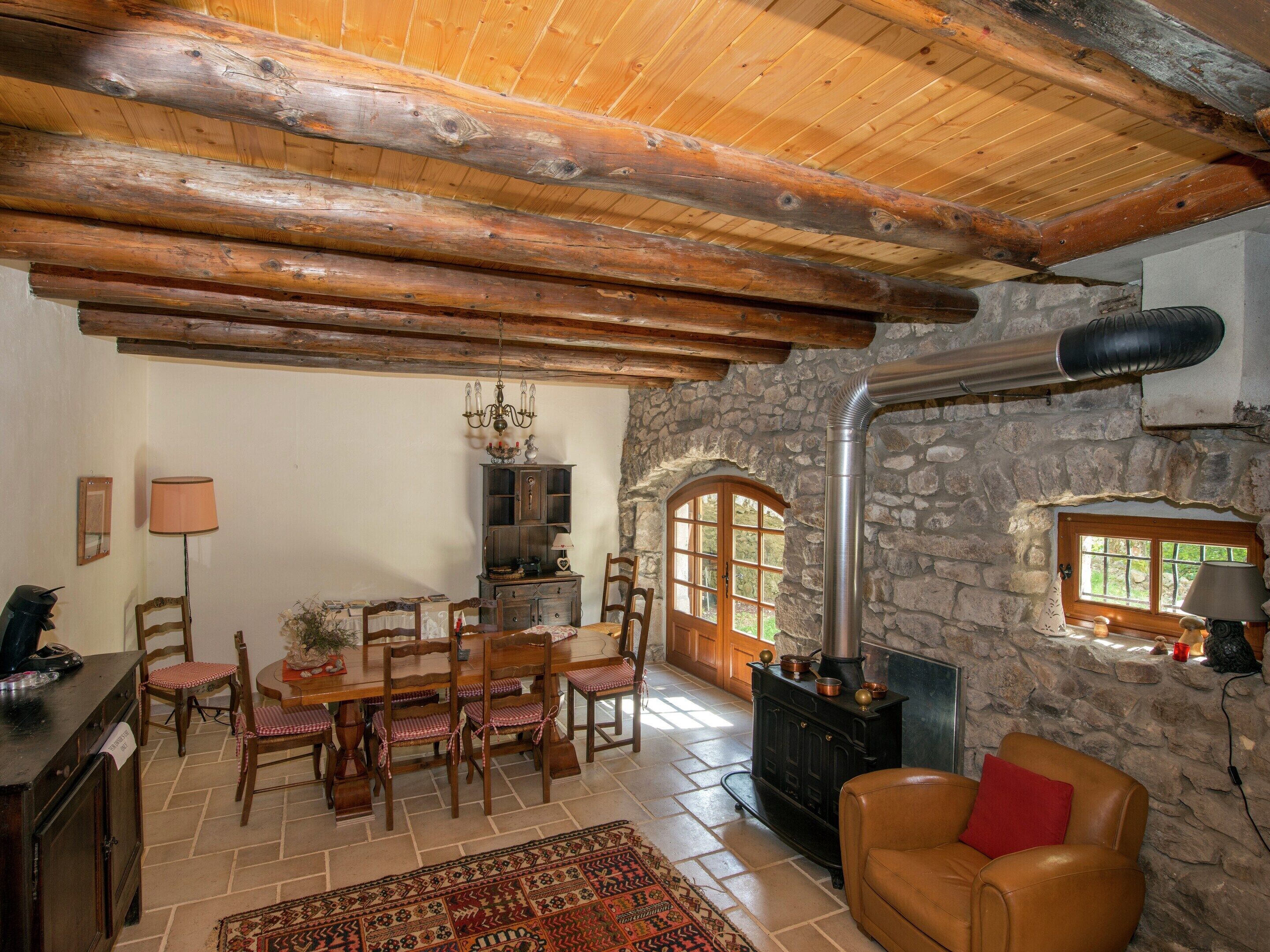 A rustic interior with a stone wall, wooden ceiling beams, a fireplace, a dining area with chairs and a table, and a leather sofa.