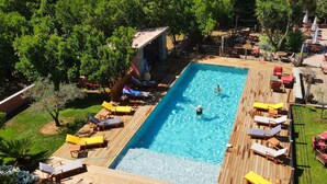 Une piscine rectangulaire dotée d’une terrasse en bois, entourée de chaises longues et de tables, et ombragée par des arbres.